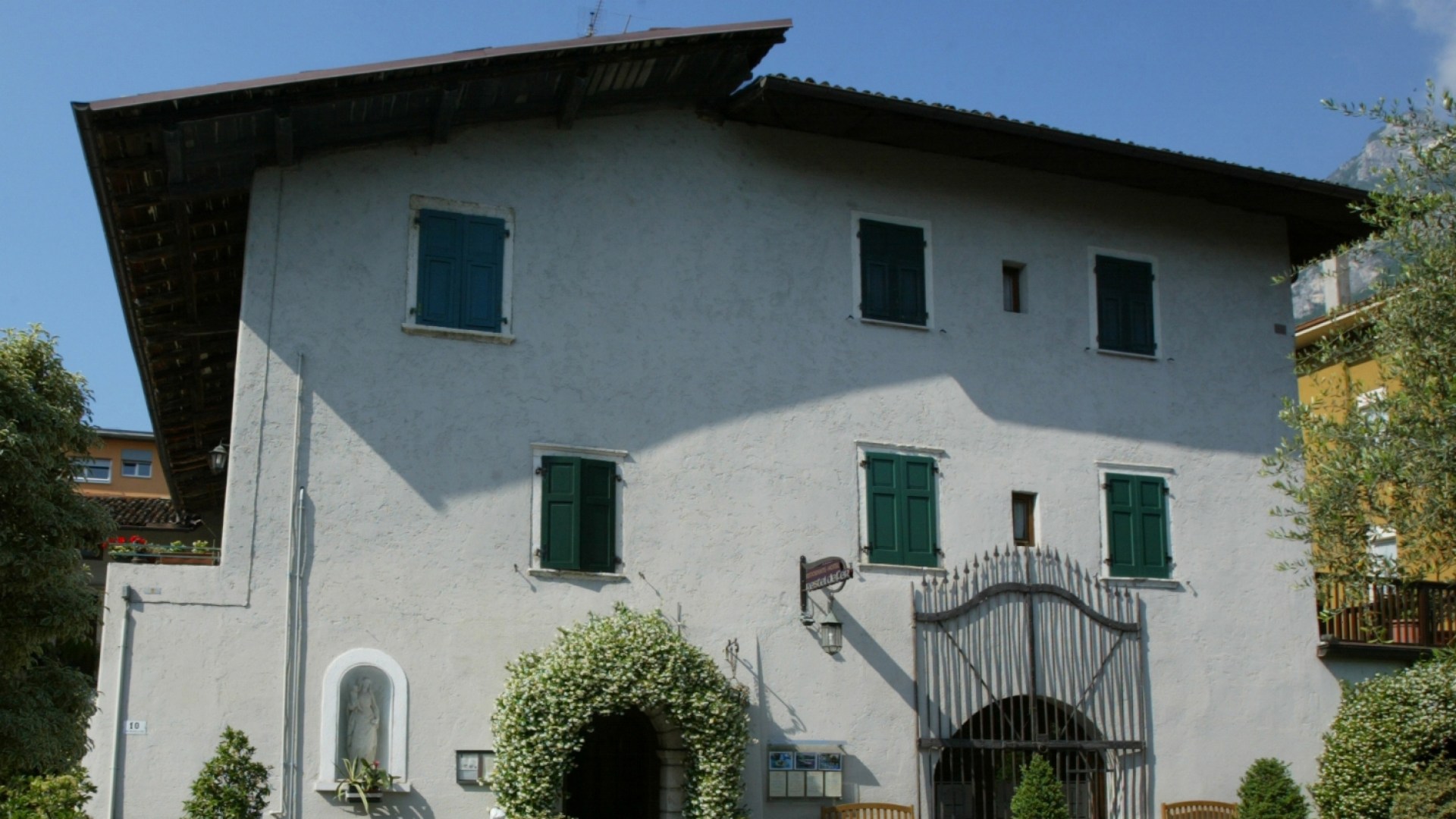 Casa tradizionale con persiane verdi, arco fiorito e statua in nicchia.