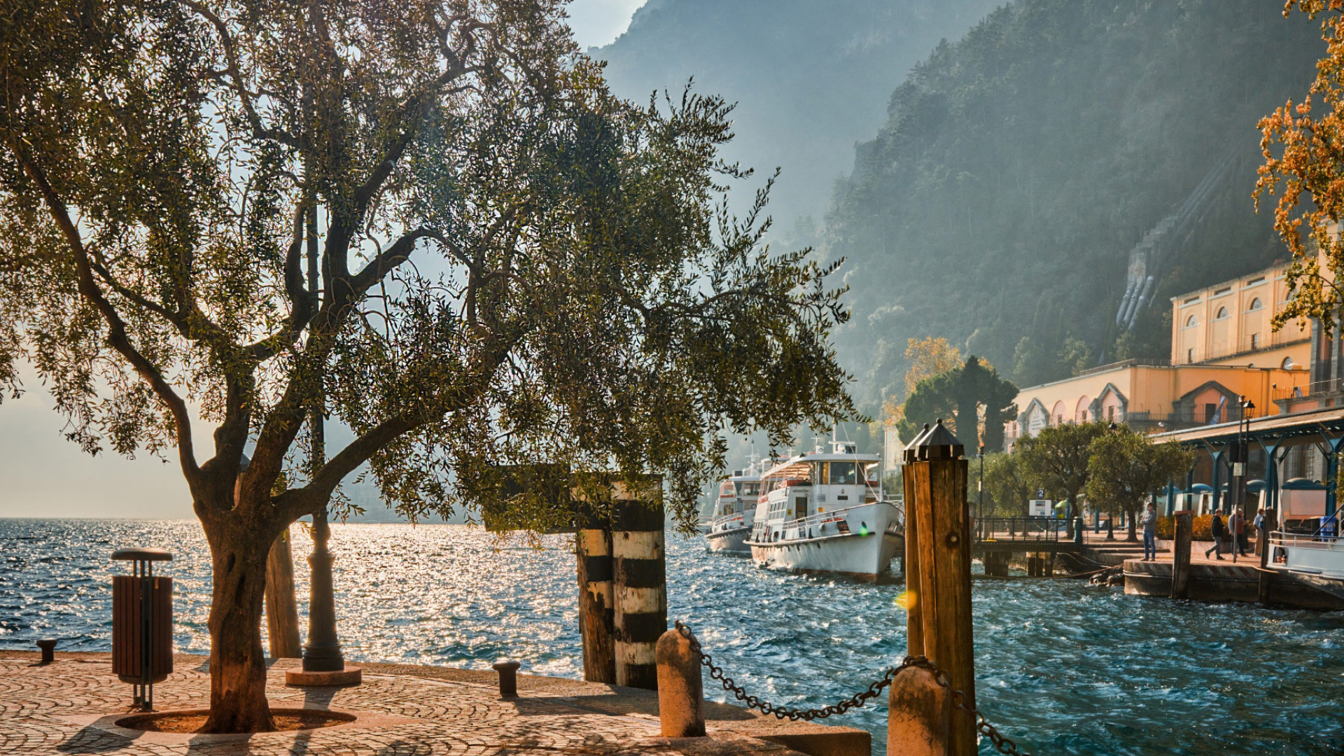 Paesaggio lacustre con barche, alberi e montagne al tramonto.