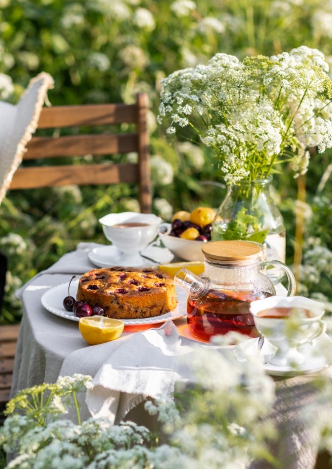 Tavolo da tè all'aperto con torta, frutta, tè e fiori bianchi.